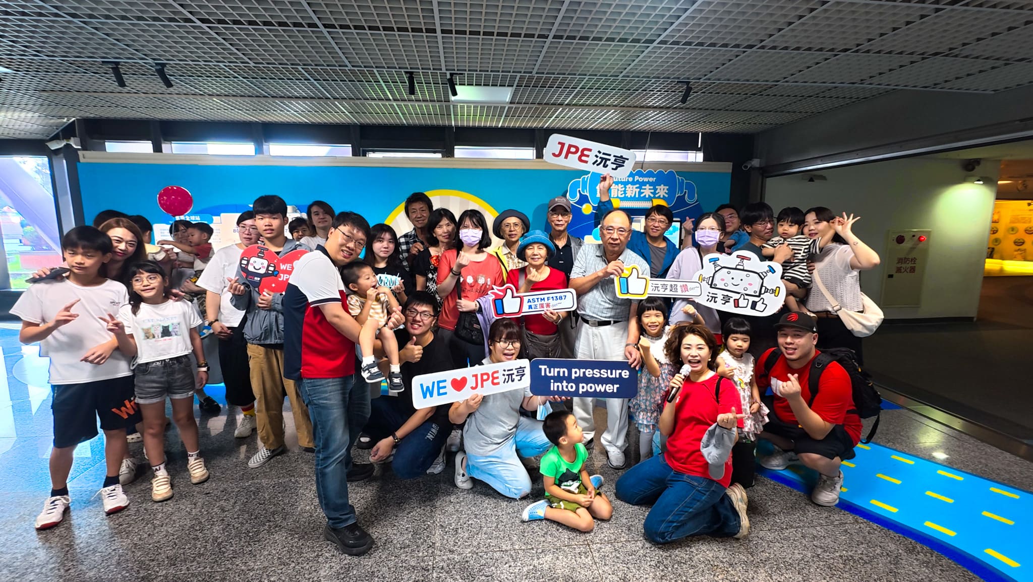 Group photo of JPE employees and their families during the Family Day at the Science Museum, holding JPE-themed signs and smiling together.
