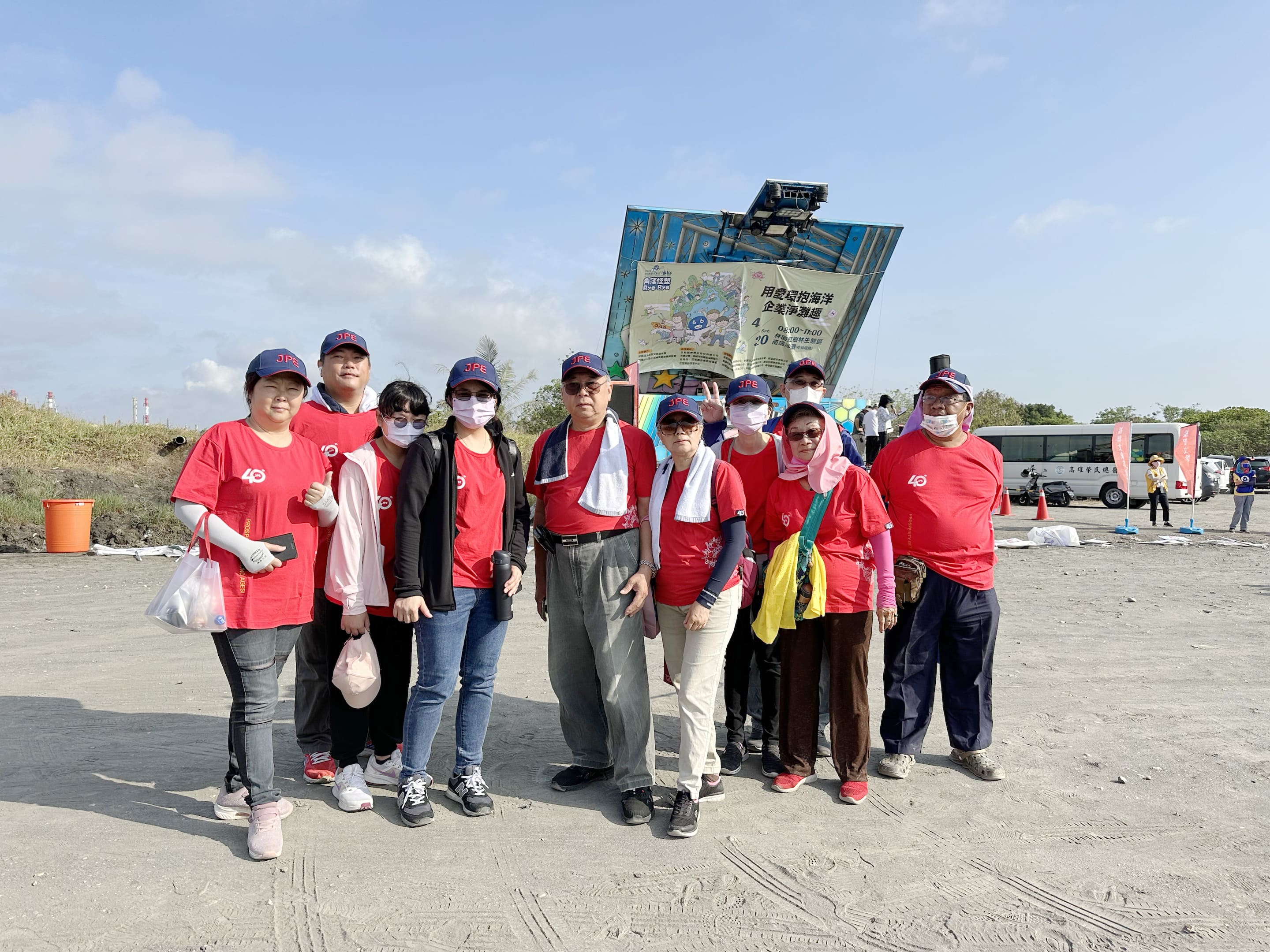 JPE Taiwan employees and family members taking a group photo at an Earth Day beach cleanup site, wearing red event shirts.