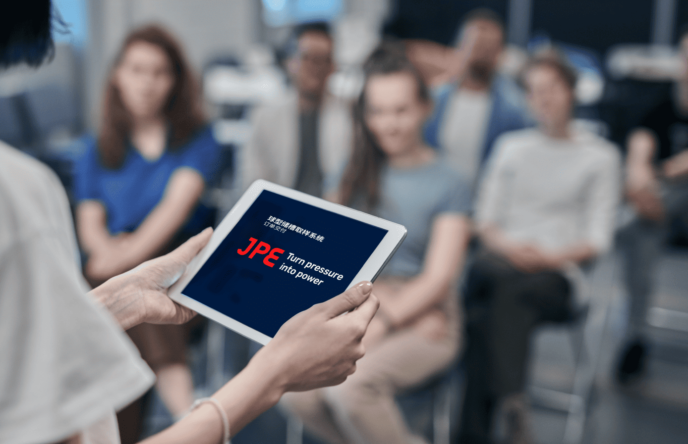 JPE technical presentation with a speaker showing sampling system information on a tablet while attendees listen attentively.
